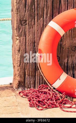 Orange lifebuoy with red rescue rope hanging on a weathered wooden post beside a swimming area. Stock Photo