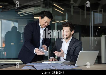 Diverse businessmen are discussing a project, collaborating on new strategies, and utilizing a digital tablet while working at an office desk with pap Stock Photo