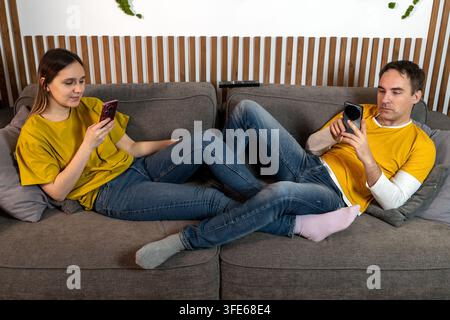 Young couple lying together on a sofa while absorbed in their smartphones, illustrating digital loneliness, emotional distance, and disconnection desp Stock Photo