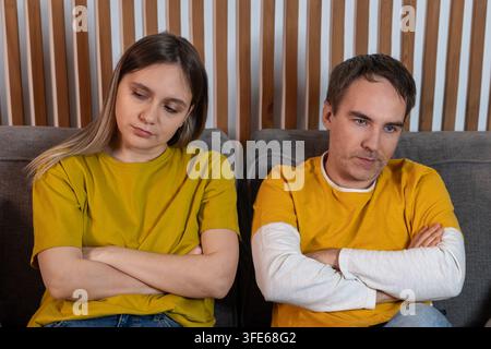 Young married couple sitting on a sofa with crossed arms, refusing to talk to each other. Family conflict, resentment, and silent treatment. Stock Photo
