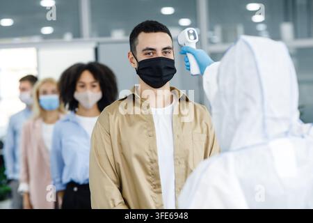 Checking temperature in morning before work. Man in hazmat suit checks first symptoms of illness in workers in protective masks in office Stock Photo