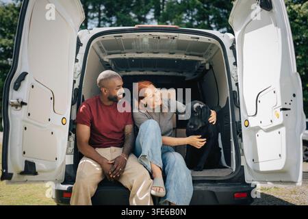 Happy diverse couple petting dog while sitting in back of moving van Stock Photo