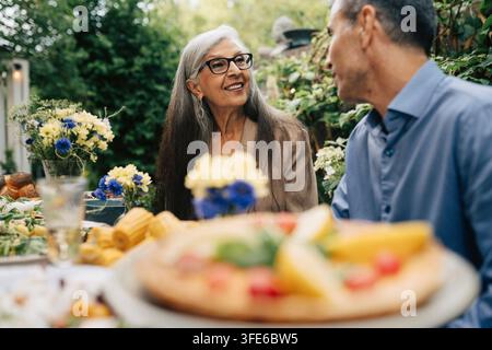 Smiling senior woman sharing conversation with man at backyard lunch party Stock Photo