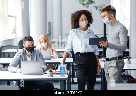 New project and plans for work. Boss in protective mask with tablet gives instructions to workers in masks, african american woman looking at gadget Stock Photo