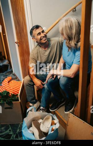 Happy man talking to woman sitting on wooden stairs in new apartment Stock Photo