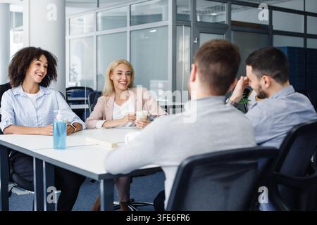 Coffee break with colleagues in rest room. Smiling multiracial colleagues with cups talking and sitting at table with antiseptic Stock Photo