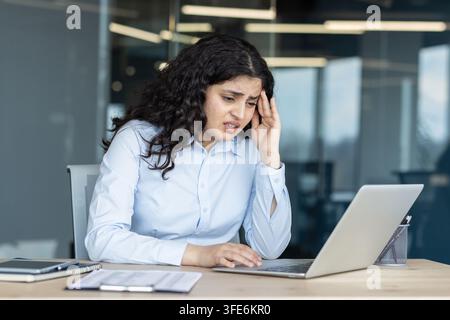 Young woman experiencing stress and frustration, holding her temple while working on a laptop, indicating an intense headache and feeling overworked in a modern office environment Stock Photo