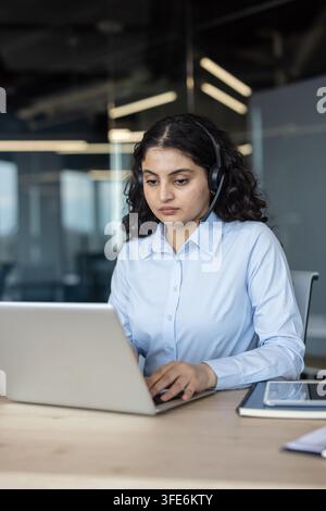 Young professional woman sitting at a desk, typing on a laptop and wearing a headset while working in an office environment, providing support and communication Stock Photo