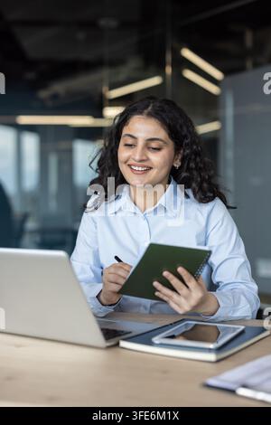Smiling young businesswoman working on a laptop while writing important information in her notebook during an online conference or study session in a modern office environment Stock Photo