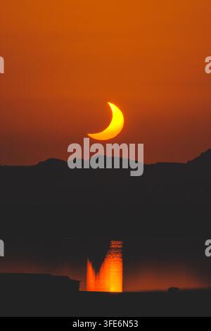 Partial solar eclipse at sunset appearing over dark mountain silhouettes with a glowing orange reflection shimmering on tranquil water, peaceful atmos Stock Photo
