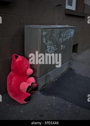 Bright pink teddy bear leans against gray utility cabinet in narrow urban alley, with weathered wall, graffiti marks, wet pavement, and muted tones cr Stock Photo