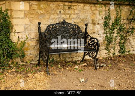 A detailed view of a black decorative cast iron bench placed against a rustic stone wall with green climbing ivy. Stock Photo