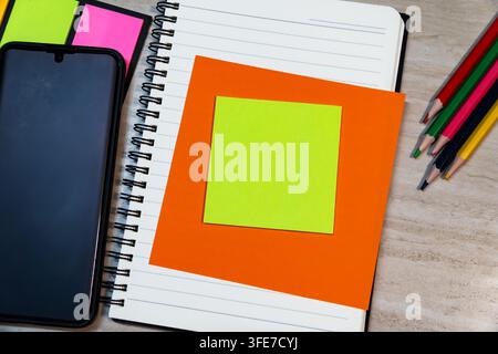 Top view of open spiral notebook with yellow sticky note on orange paper, smartphone, and colored pencils on wooden desk background. Stock Photo