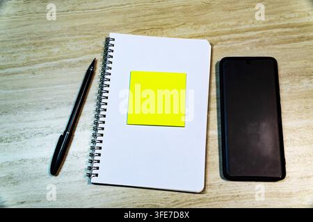 Top view of open spiral notebook with yellow sticky note, pen, and smartphone on wooden desk. Copy space for mockup or text. Stock Photo