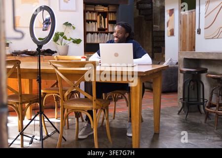 African American man filming smartphone in ring light at dining table using laptop wearing navy top Stock Photo