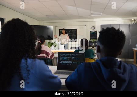 Female teacher with teens gesturing and teaching in classroom with laptop showing code in sweater Stock Photo