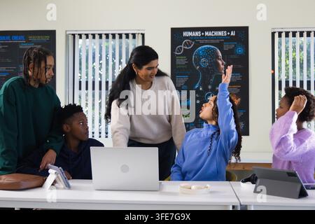 Indian teacher leaning in engaging African American teenage students at classroom table with laptop Stock Photo