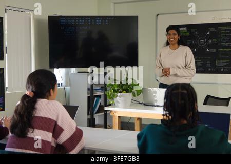 Female teacher addressing teen students in classroom with laptop, monitor with code, copy space Stock Photo
