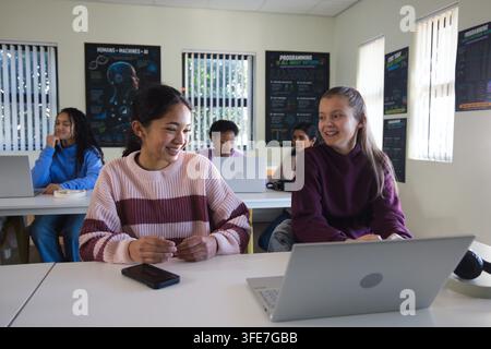 Female students smiling and talking while sitting at classroom table with laptops and smartphone Stock Photo