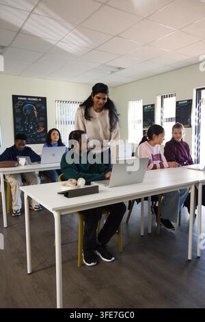 Indian woman instructor leaning over student helping with laptops headset at front table in-class Stock Photo