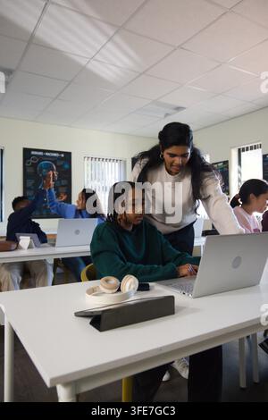 Indian teacher leaning over African American teen student, guiding on silver laptop in school Stock Photo