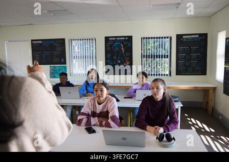 Diverse teenage students using laptops at white tables in classroom while teacher gesturing Stock Photo