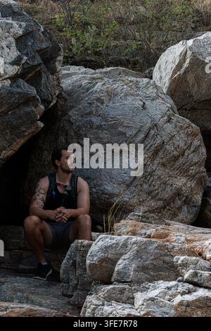 Southern Arizona - July 2022: A man sits in the shade of a rock crevice, surrounded by large weathered boulders, gazing off to the side. Stock Photo
