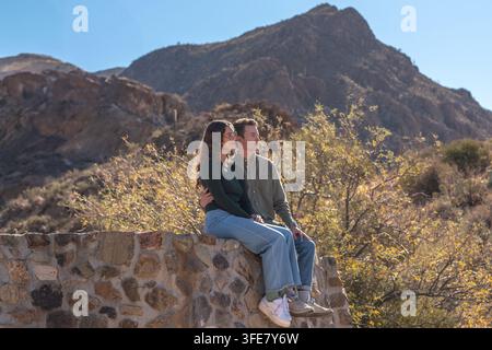 Autumn 2024: A couple sits close together on a stone wall admiring desert mountains and shrubland at sunset. Stock Photo