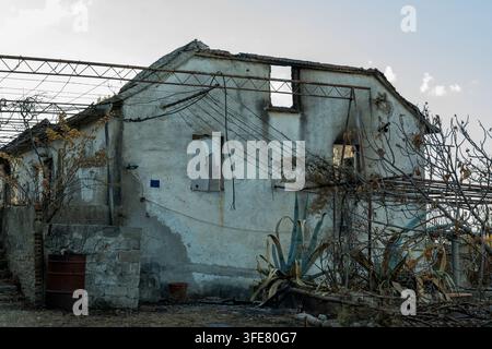 An abandoned rural house with cracked plaster, broken windows, tangled wires, and dry overgrown vegetation stands under a bright sky. Stock Photo