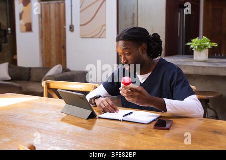 African American man in navy scrubs examining pill bottle while using tablet at home dining table Stock Photo