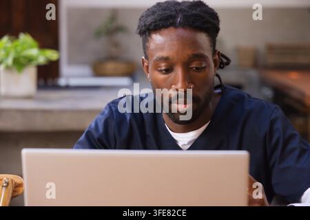 African American man working on silver laptop in home kitchen wearing navy scrub top Stock Photo
