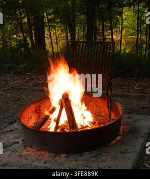 Bright flames rising from a campfire in a fire ring when the campsite is dark, but the lake behind is still bathed in light. Stock Photo