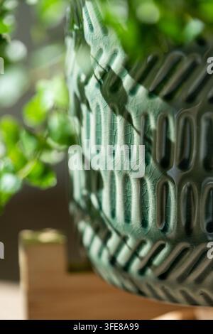 Close-up of textured green ceramic plant pot with blurred foliage in soft natural sunlight. Stock Photo