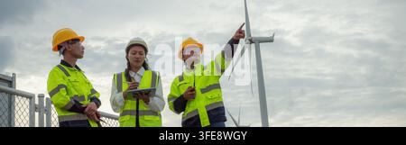 Team of Asian engineers holding digital tablet and pointing up while inspecting wind turbine power plant site. Diverse male and female technicians wea Stock Photo