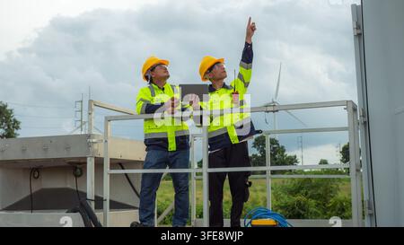 Two Asian male engineers holding digital tablet and pointing up while inspecting wind turbine power plant site. Electrical technicians wearing hardhat Stock Photo