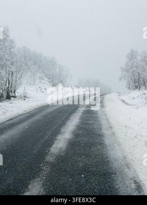 A cold and atmospheric winter landscape in Norway, showing a wet, dark asphalt road curves gently through a snow-covered environment Stock Photo