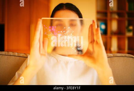 Young woman interacts with a transparent screen showing digital connections while seated on a couch in a contemporary living room with wooden shelves Stock Photo