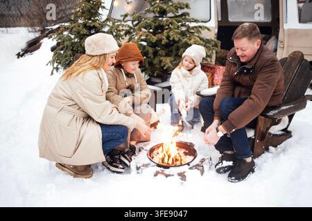Family enjoys roasting marshmallows around a campfire in winter, seated on wooden chairs with snow-covered ground and evergreen trees surrounding them Stock Photo