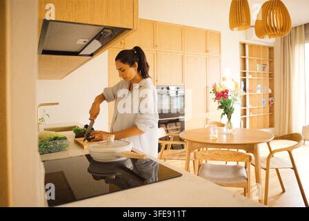 Woman in light blue shirt prepares vegetables on a wooden cutting board in a contemporary kitchen, featuring wooden cabinets and a dining area Stock Photo