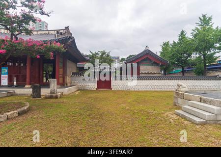Traditional Korean buildings with ornate tiled roofs, red columns, stone walls, monuments, and flowering trees stand against a modern urban backdrop under an overcast sky. Stock Photo