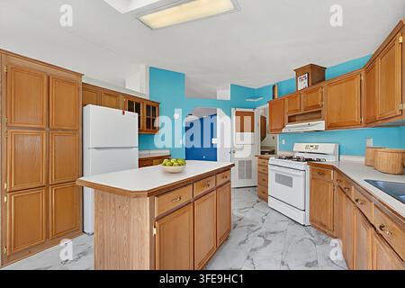 Bright blue kitchen scene with warm maple cabinets, white refrigerator and stove, kitchen island, skylight delivering clear daylight, tiled floor for Stock Photo