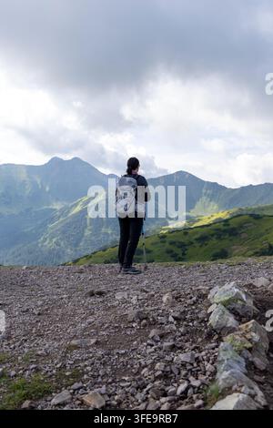 Female hiker with backpack standing on rocky ridge looking at lush green mountain valley. Stock Photo