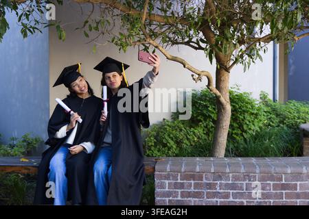 Diverse female grads sitting on campus bench in black gowns holding diplomas and cell taking selfie Stock Photo