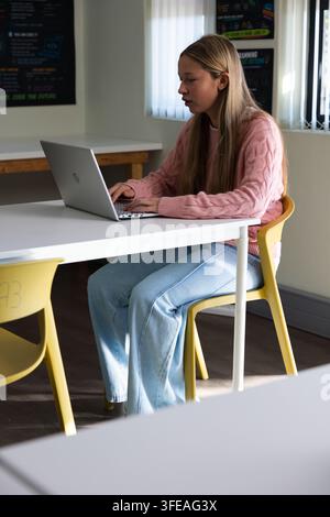 Teenage female wearing pink sweater and light blue jeans, typing on silver laptop at white table Stock Photo