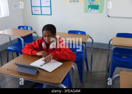 African teenage student taking notes in classroom using tablet, in red hoodie and pink headphones Stock Photo