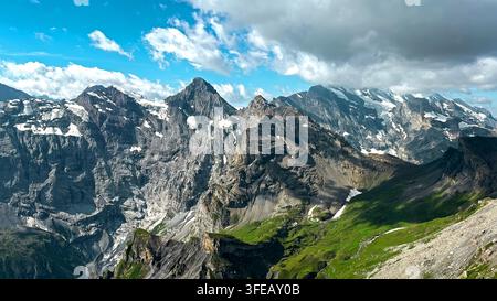 Dramatic Snowcapped Alpine Mountain Range Under Clouds. Rugged snowcapped mountain peaks rise above a green alpine valley beneath a dramatic blue sky Stock Photo