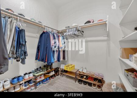 Spacious walk-in closet featuring hanging clothes along two walls, a bench, shelves with hats and boxes, and a floor with sneakers and dress shoes und Stock Photo