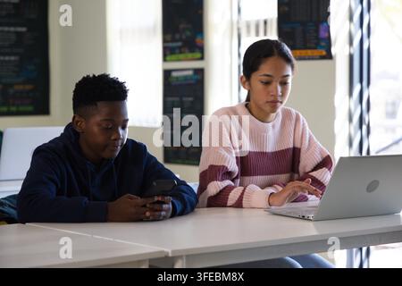 Diverse teens sitting in classroom at table in hoodie and pink sweater, using smartphone and laptop Stock Photo