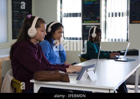 Diverse youth students wearing hoodies and white headphones using laptops and tablet in classroom Stock Photo