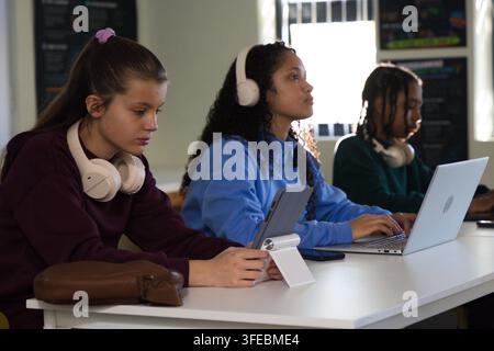 Diverse teenage students sitting at white table in classroom, using laptops and wearing headphones Stock Photo